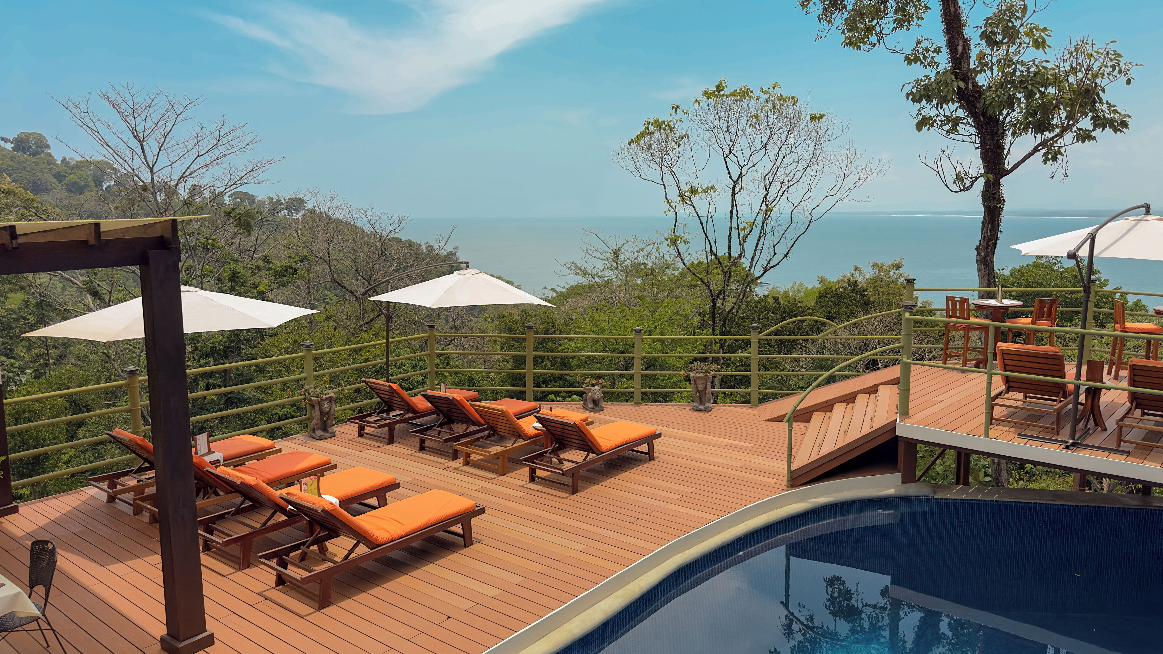 A wooden deck with lounge chairs and umbrellas overlooking a pool and a scenic view.