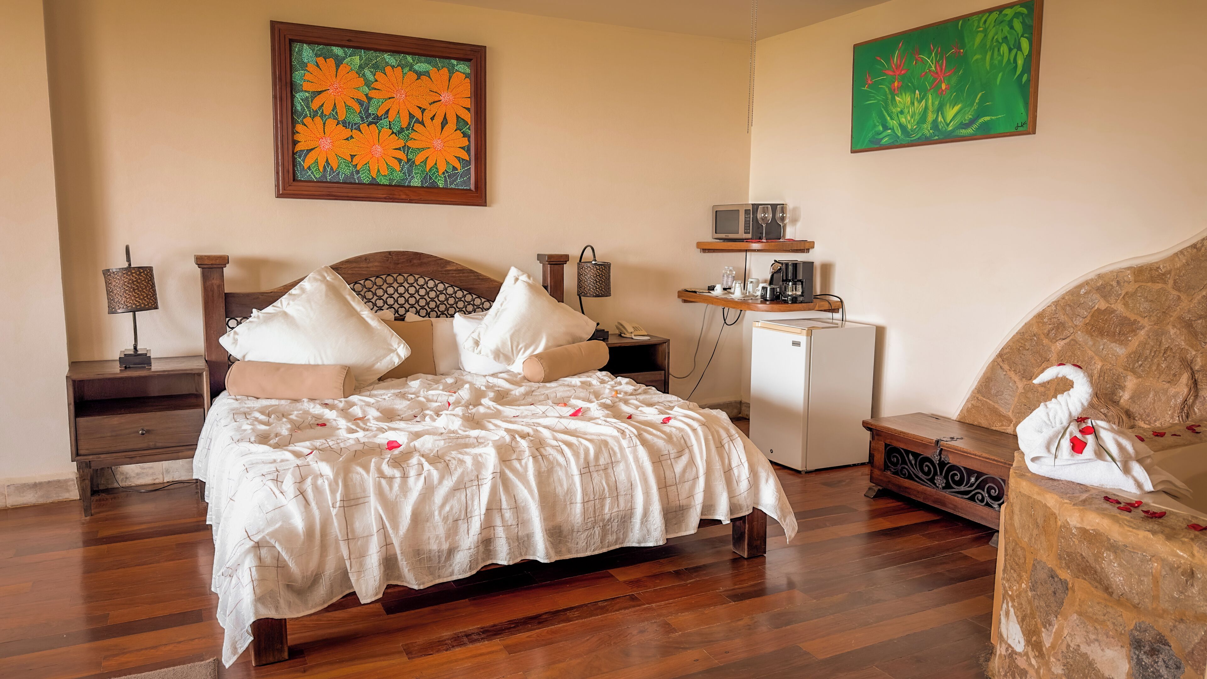 A bedroom with a bed, bedside tables, a small refrigerator, and artwork on the walls.