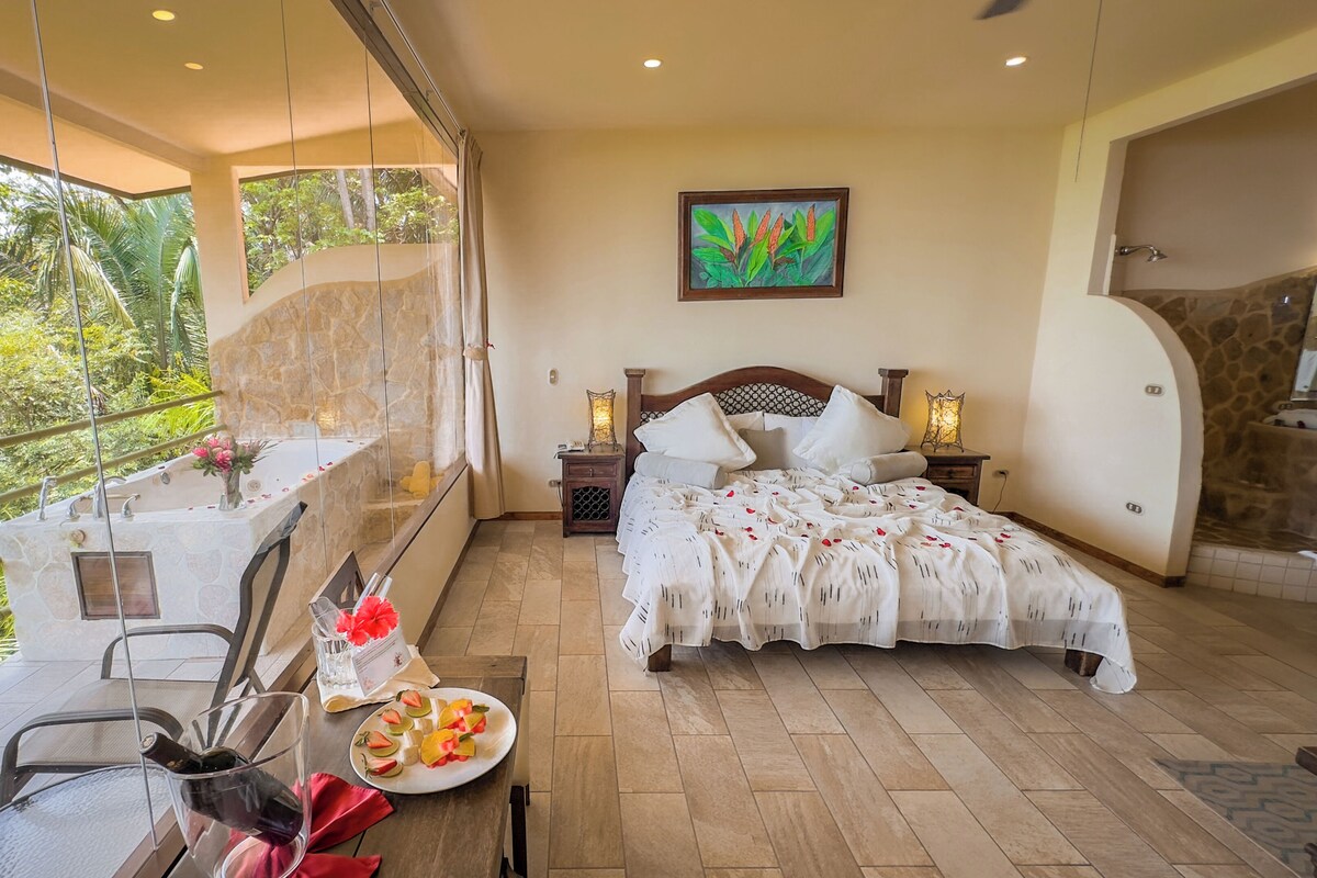 A bedroom with a large bed, a bedside table, a nightstand, a window with a view of greenery, and a balcony with a table and chairs.