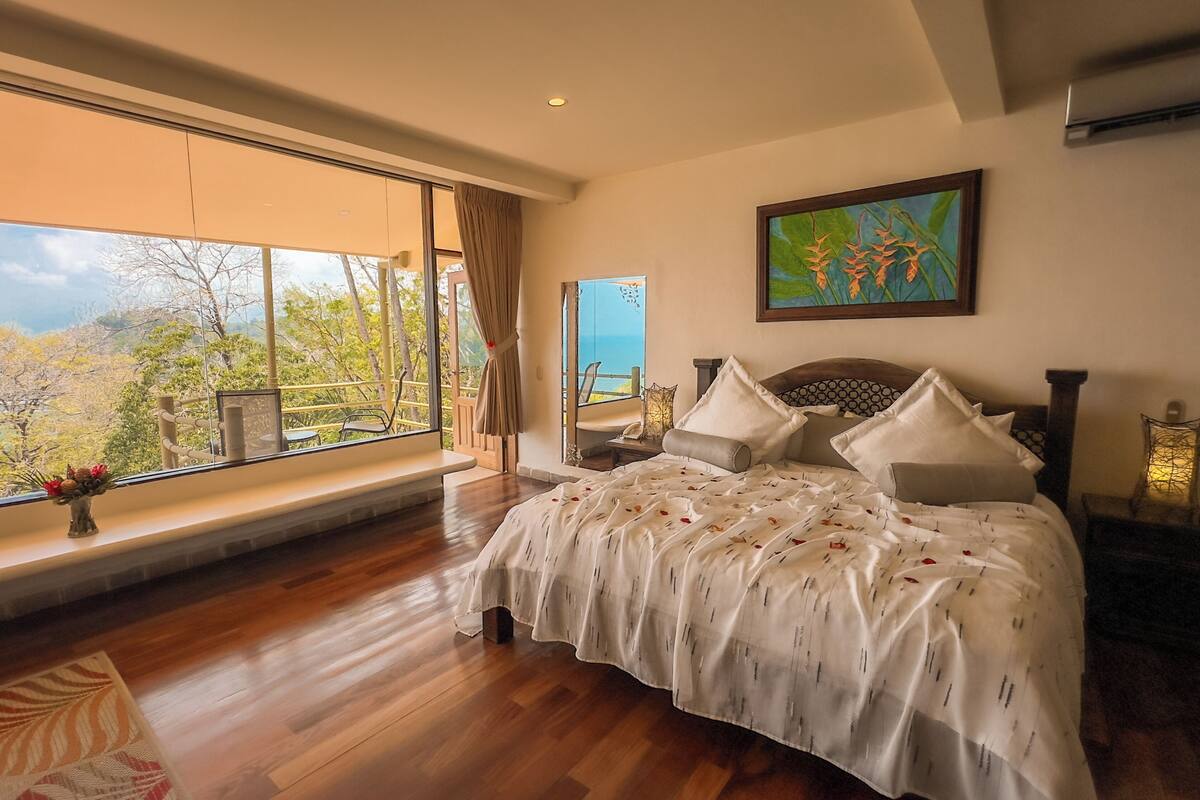 A bedroom with a large window, a bed with white bedding, a wooden floor, and a painting on the wall.