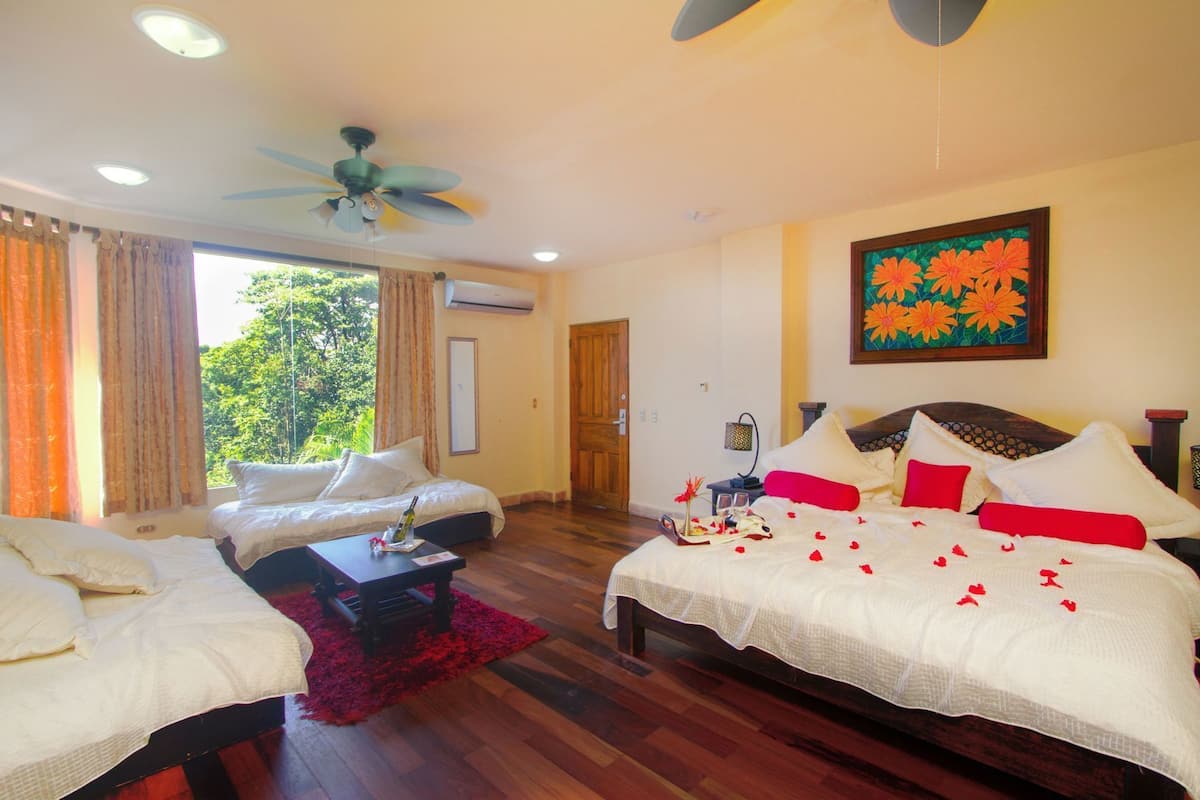 A hotel room with two beds, a ceiling fan, a painting, and a view of greenery outside.