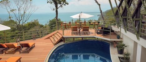 A pool area with wooden deck loungers, a small pool, and a view of the sea.