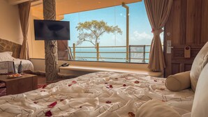 A hotel room with a large bed, a TV, a balcony with a view of trees and the sea, and a wooden desk with a bottle and glasses.