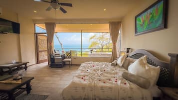 A bedroom with a large bed, a desk, and a chair, a ceiling fan, and a view of the sea.
