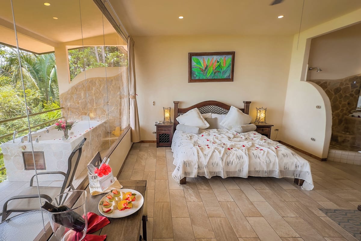 A bedroom with a large bed, a bedside table, a nightstand, a window with a view of greenery, and a balcony with a table and chairs.