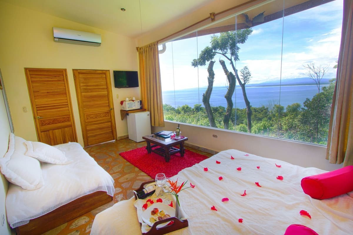 A hotel room with two beds, a TV, a small kitchenette, and a large window with a view of trees and water.