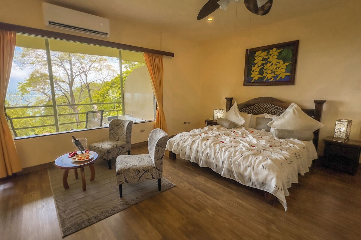 A bedroom with a large window, a bed with white bedding, a chair, a small table with a bottle, and a decorative wall piece.