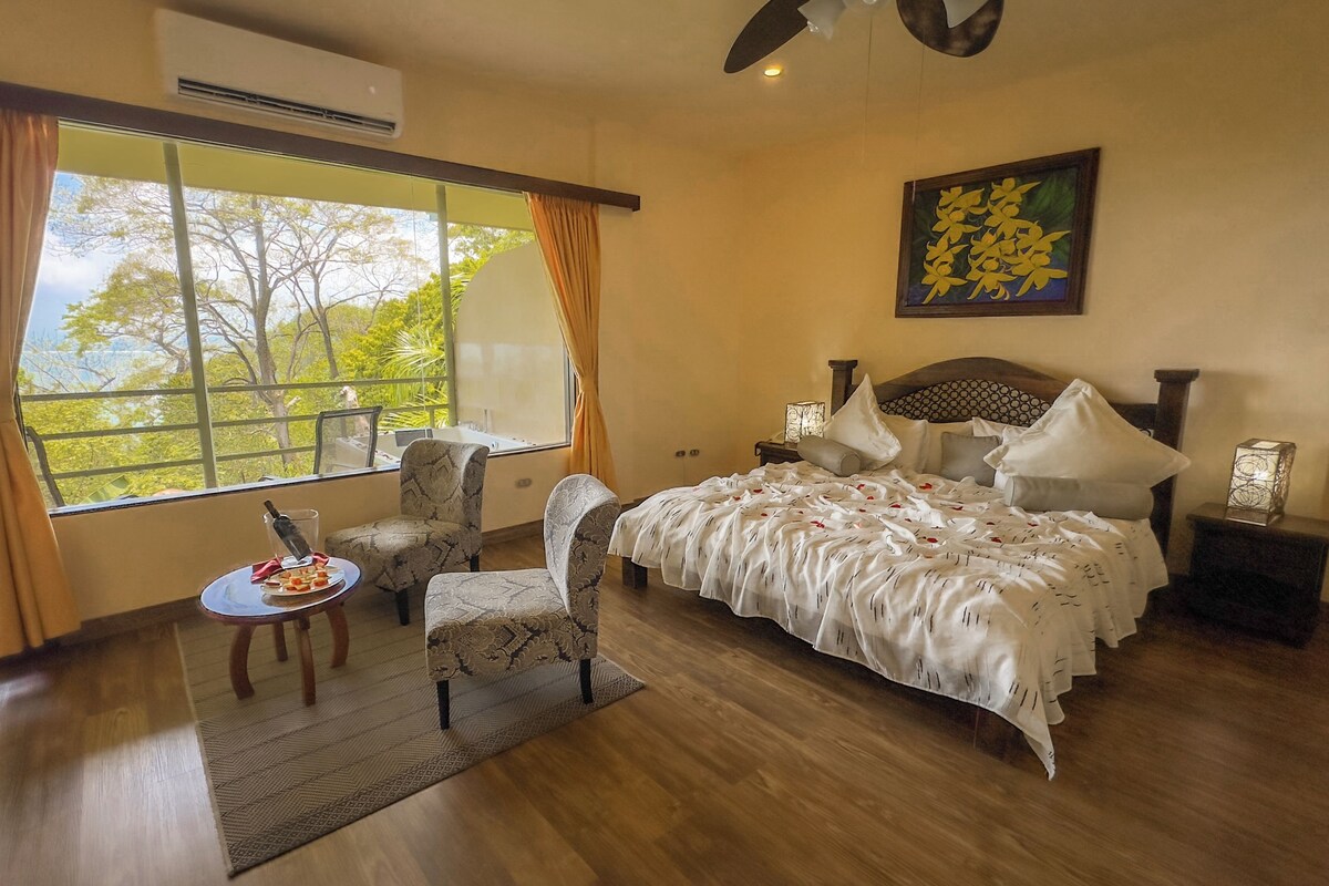 A bedroom with a large window, a bed with white bedding, a chair, a small table with a bottle, and a decorative wall piece.