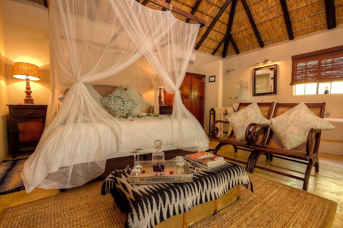 A bed with a canopy, a bedside table with a lamp, a wooden bench with cushions, and a tray with glasses and a book.