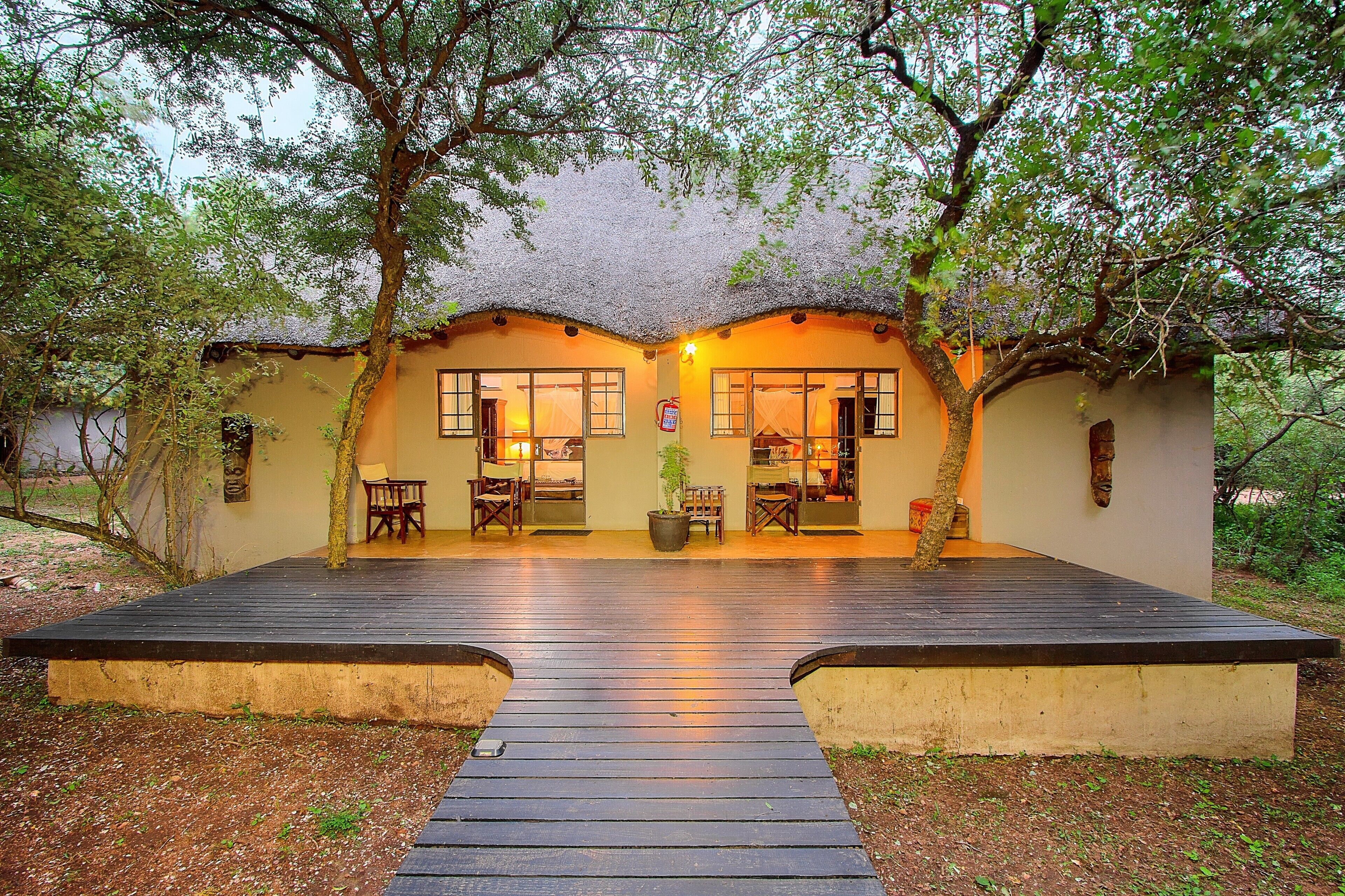 A cozy lodge with a wooden deck, two seating areas with chairs, and a thatched roof.