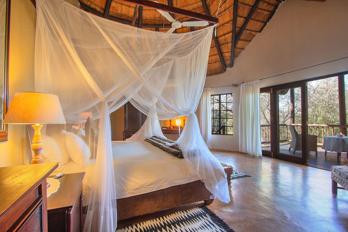 A bedroom with a four-poster bed, a nightstand, a lamp, and a view of the outdoors through large windows.