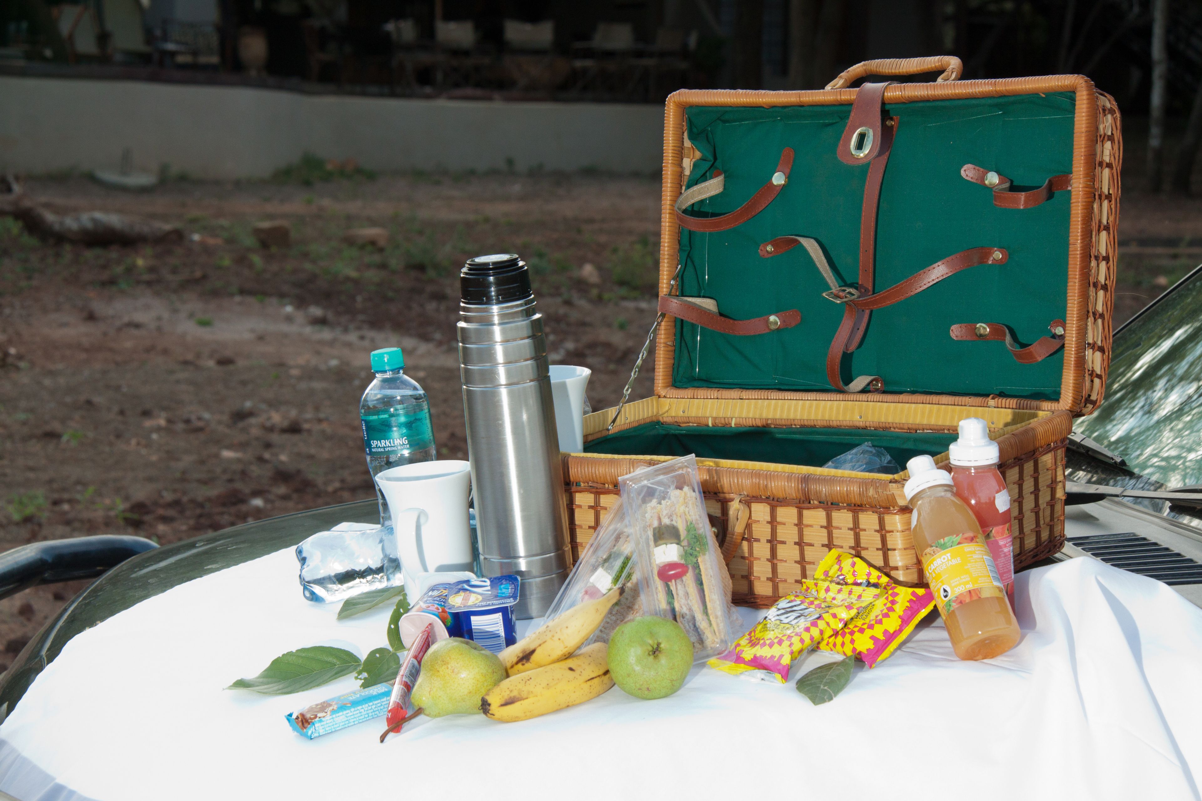 A picnic setup with a wicker basket, food items, a thermos, and a bottle on a table.