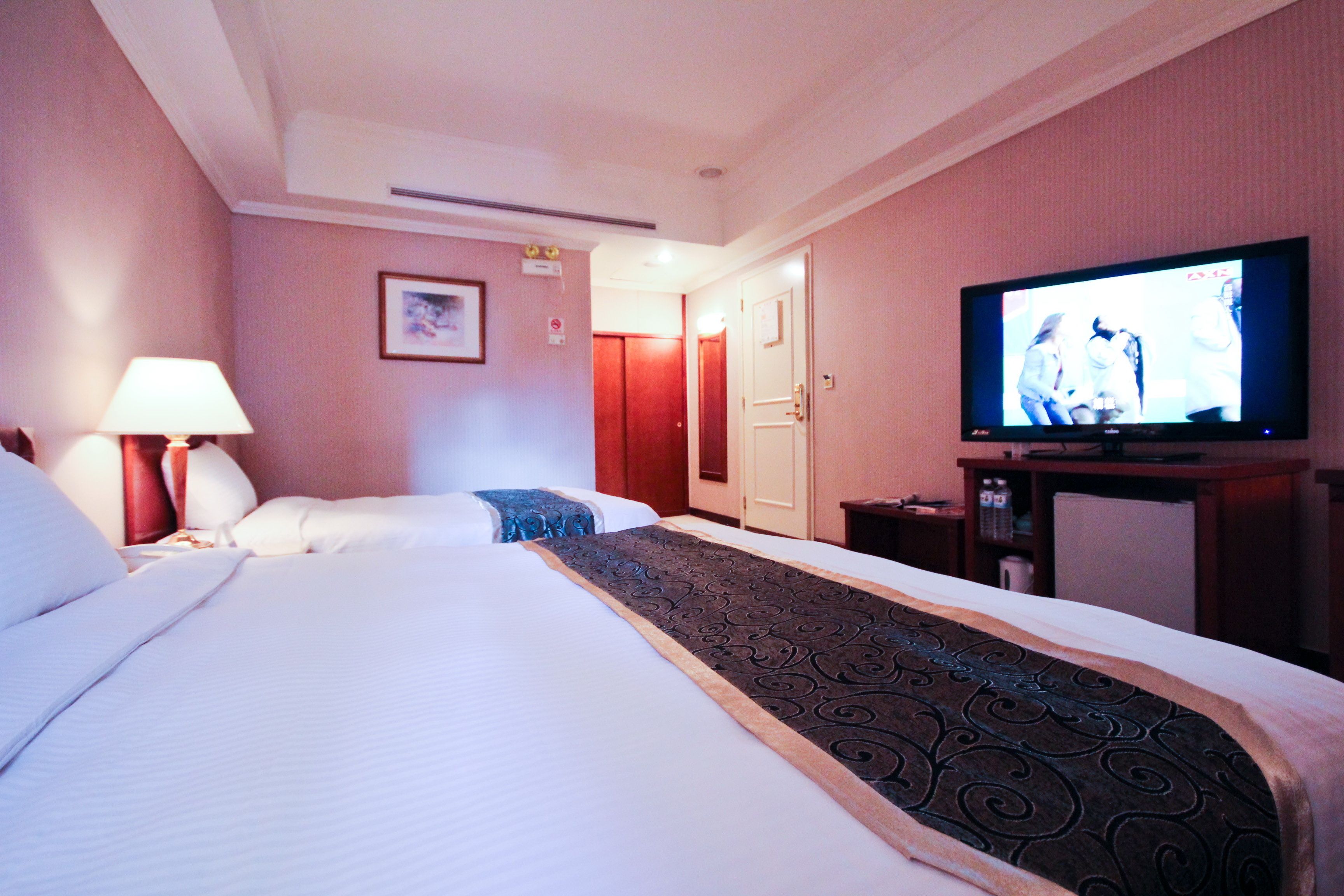 A hotel room with two beds, a television, a lamp, and a door leading to another room.