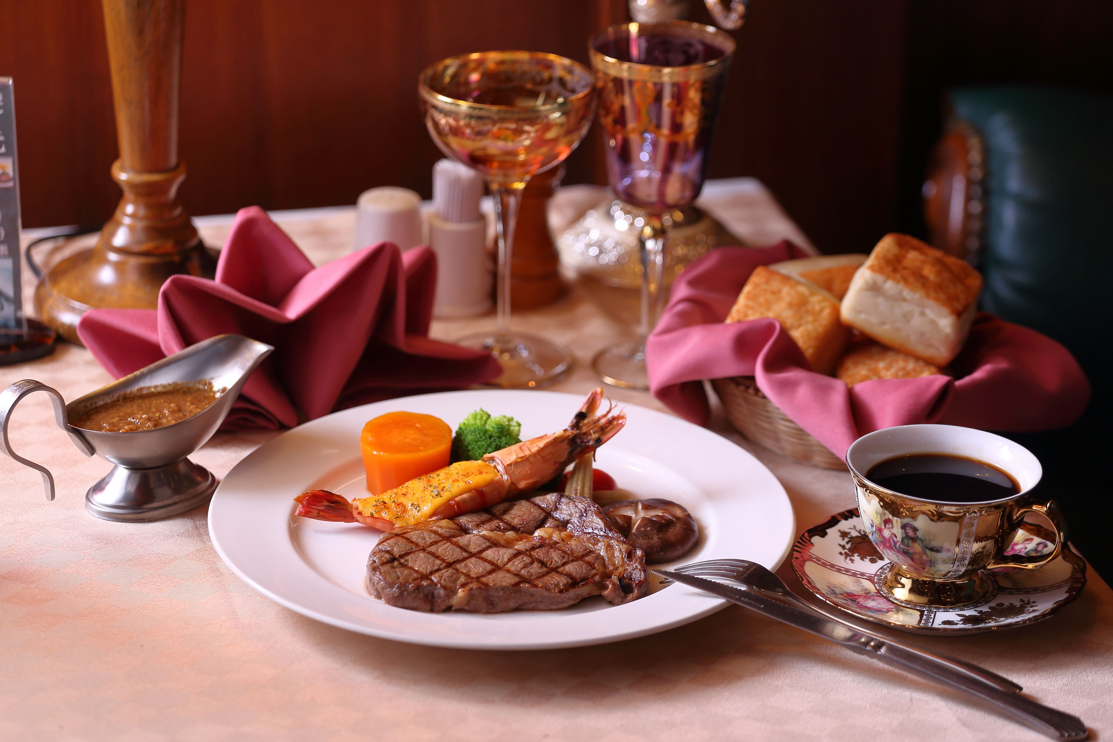 A plate with steak, lobster, and vegetables, a cup of coffee, and a basket of bread on a table.