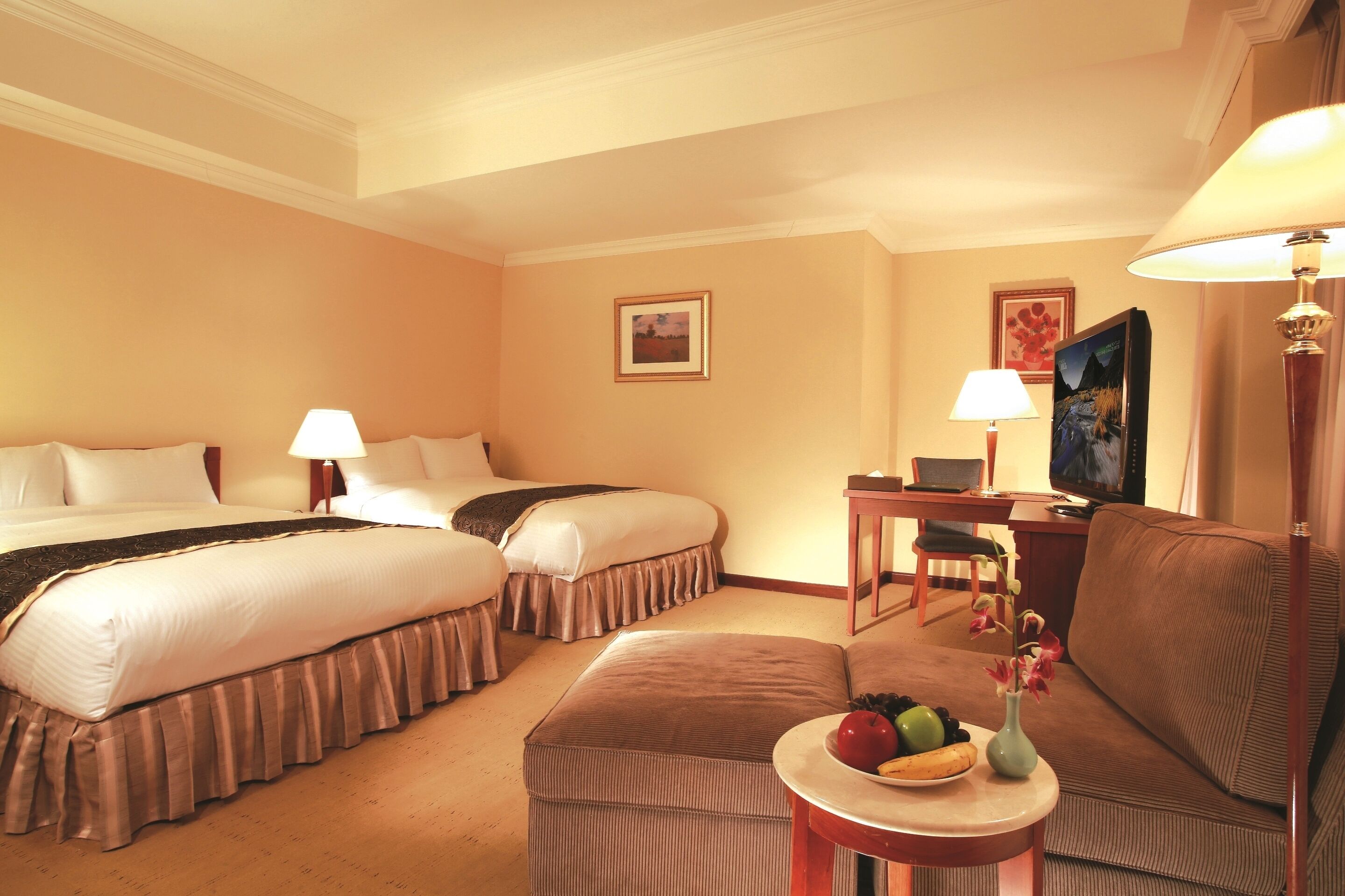 Une chambre d’hôtel avec deux lits, un bureau, une chaise, un canapé et une petite table sur laquelle sont disposés des fruits et des fleurs.
