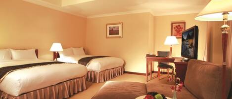 A hotel room with two beds, a desk, a chair, a sofa, and a small table with fruit and flowers.