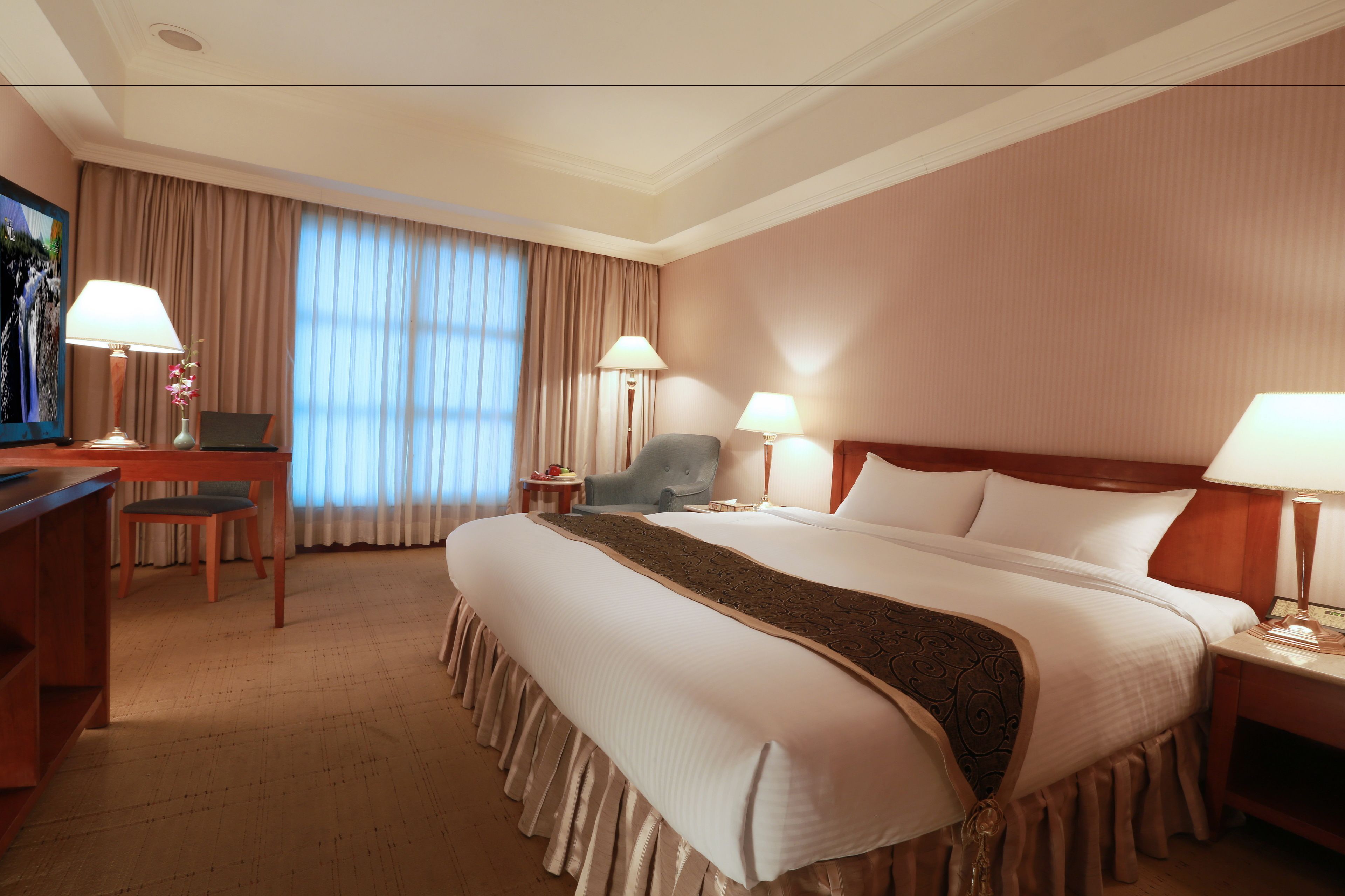 A hotel room with a large bed, a desk with a chair, a television, and two lamps.