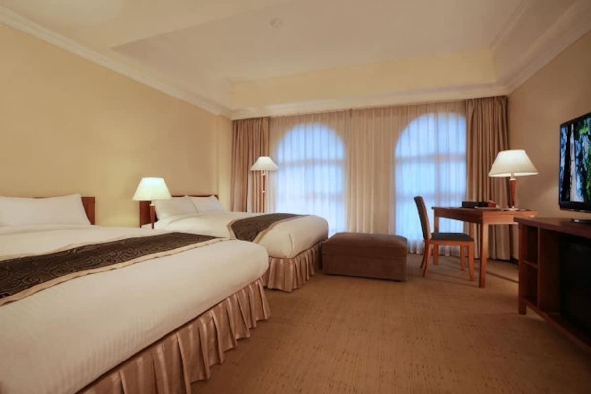 Habitación de hotel con dos camas, un escritorio, una silla, televisión y ventana con cortinas.