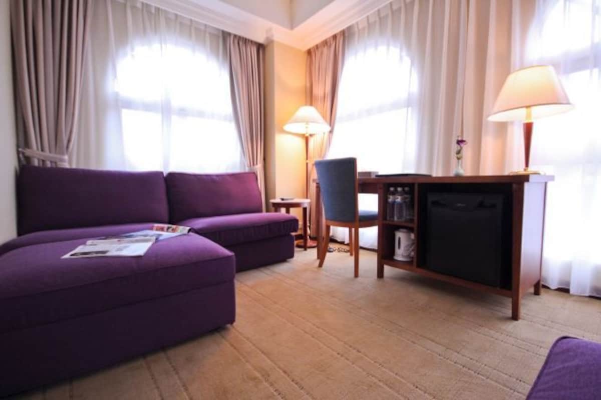 Una habitación con un sofá púrpura, una mesita pequeña, una silla, una lámpara y un mini bar.