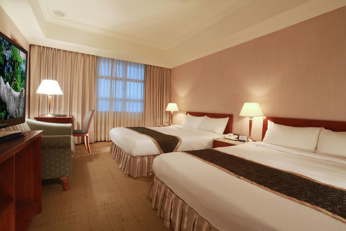 A hotel room with two beds, a desk, a TV, and a window with curtains.