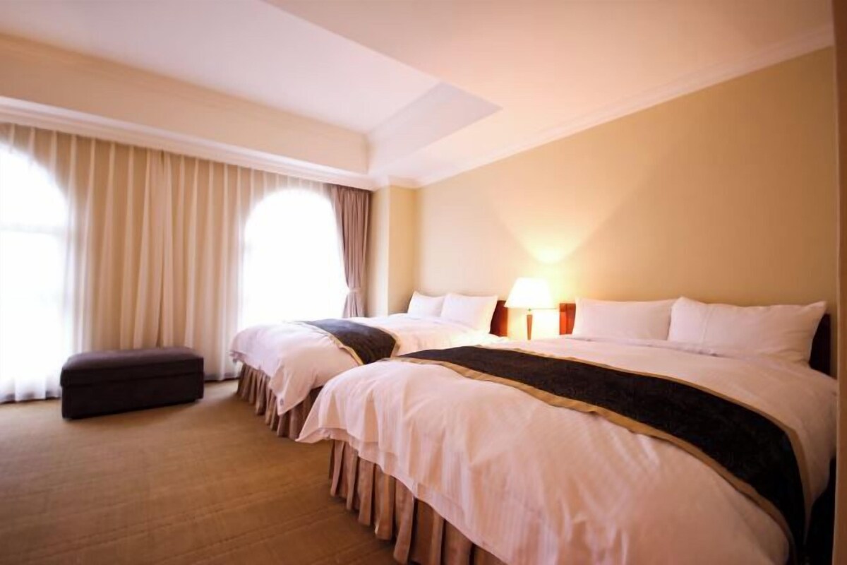Habitación de hotel con dos camas, un ventanal con cortinas, una mesita de noche con lámpara y piso alfombrado.