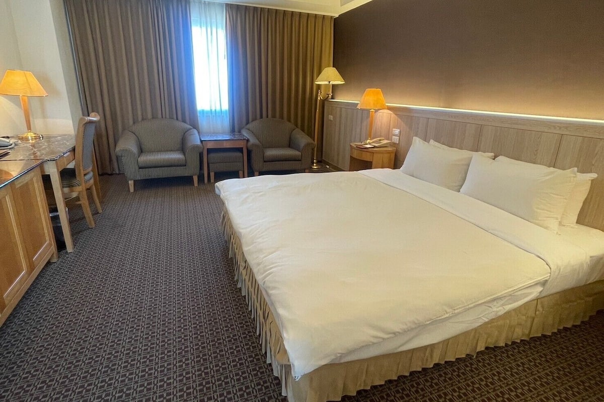 A hotel room with a large bed, two armchairs, a small table, a desk, and a nightstand.