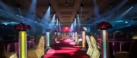 A banquet hall with a red carpet aisle, illuminated by spotlights, and flanked by tall, decorative columns with floral arrangements.