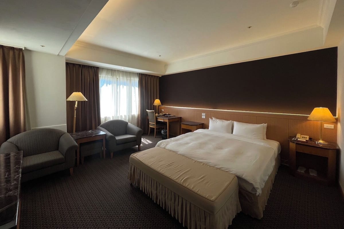 A hotel room with a large bed, a desk, a chair, a small table, and a lamp.