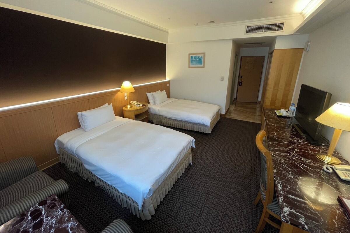 A hotel room with two beds, a desk with a TV, a chair, a sofa, and a carpeted floor.