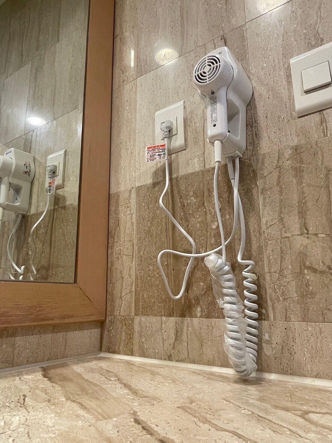 Ένα πιστολάκι μαλλιών τοποθετημένο στον τοίχο, με έναν καθρέφτη που αντανακλά την εικόνα, μια πρίζα και ένα καλώδιο τυλιγμένο σε σπείρα.