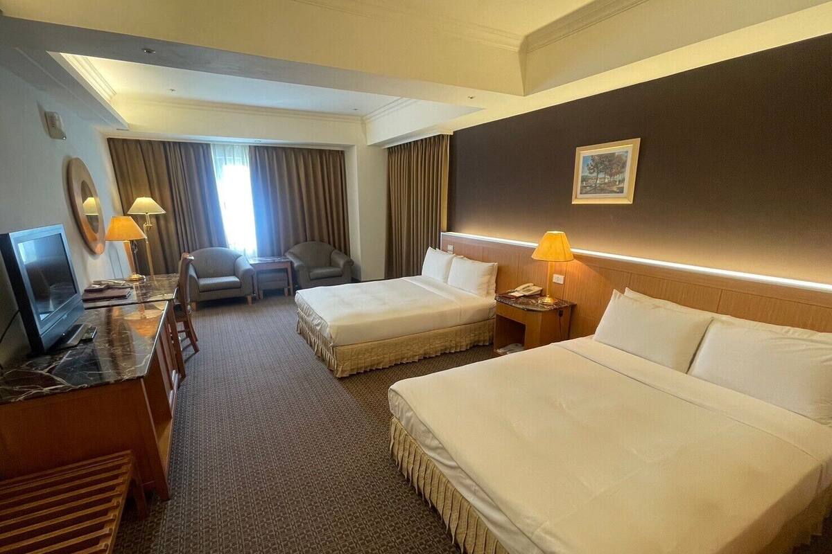 A hotel room with two beds, a desk with a TV, a chair, a lamp, and a painting on the wall.