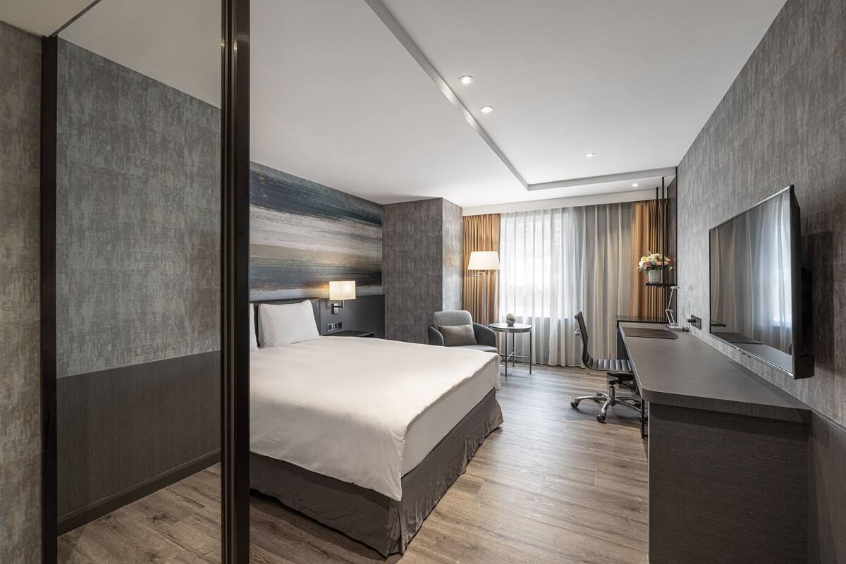 A modern hotel room with a large bed, a desk with a chair, a flat-screen TV, and a small table with a lamp.
