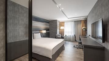 A modern hotel room with a large bed, a desk with a chair, a flat-screen TV, and a small table with a lamp.