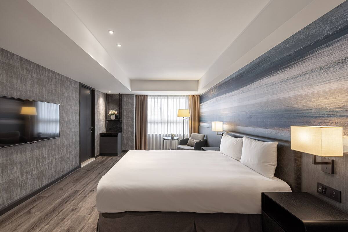 A modern hotel room with a large bed, a flat-screen TV, a small table, and a seating area with a lamp.
