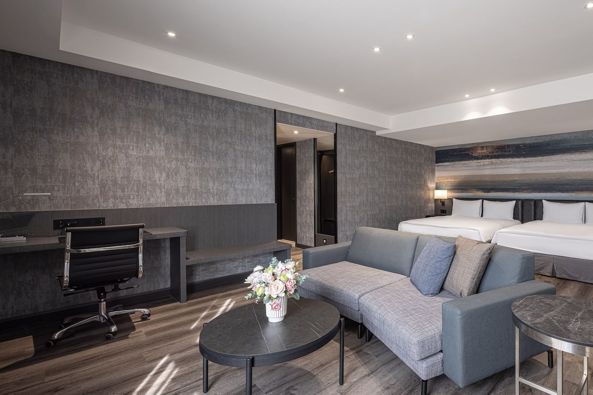 A modern hotel room with a grey sofa, a small round table, a desk with a chair, and two beds with headboards.