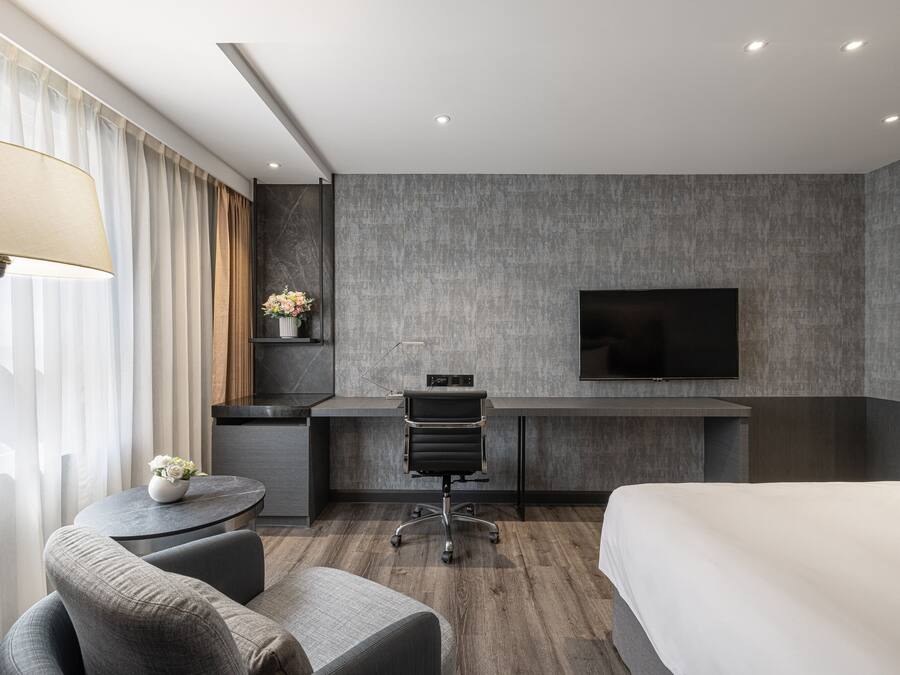 A modern hotel room with a bed, a desk with a chair, a TV, and a small table with a vase of flowers.