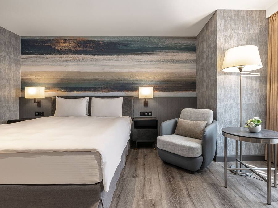 Une chambre d’hôtel moderne avec un grand lit, un petit fauteuil, une table d’appoint avec une lampe et une table de chevet fixée au mur.