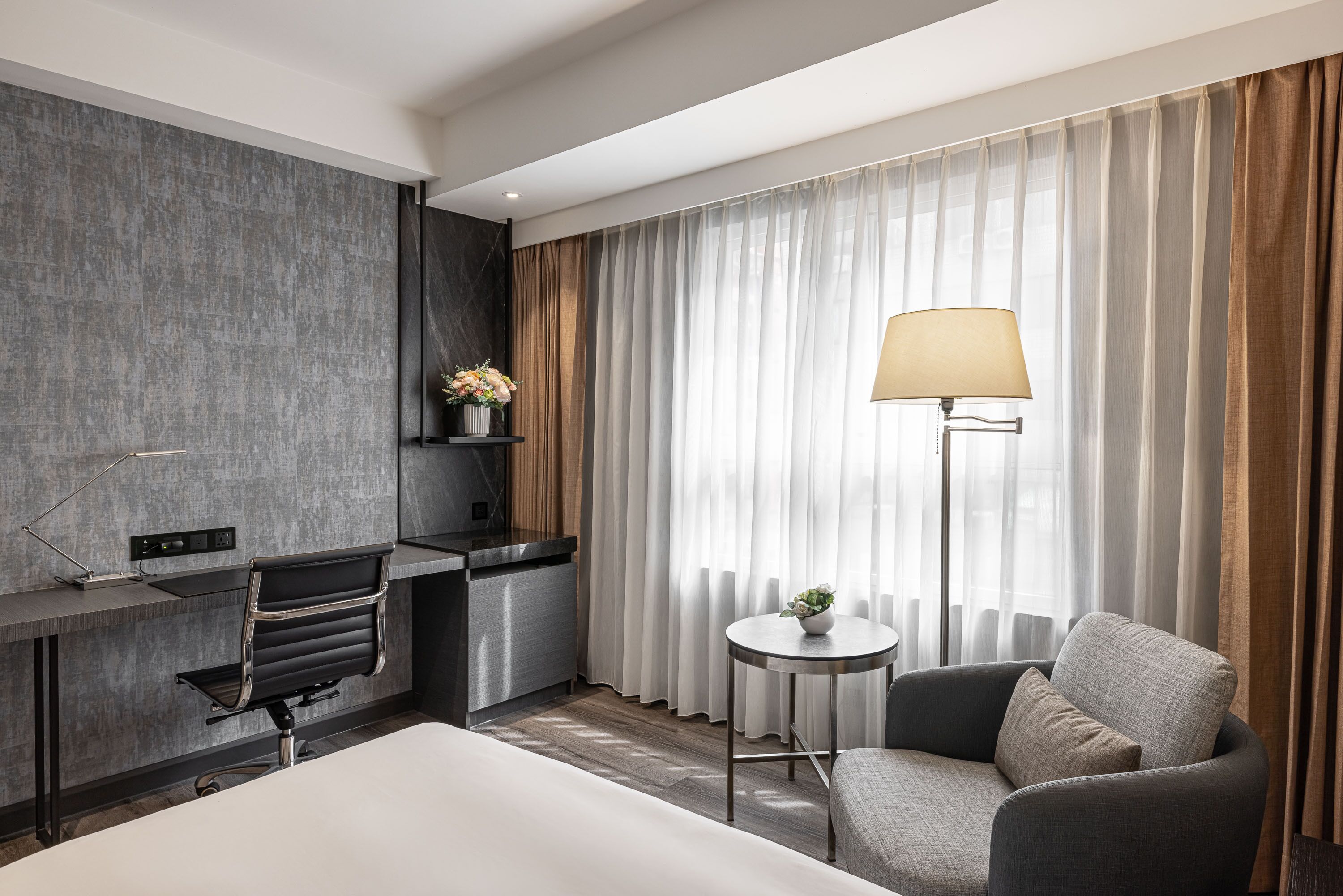 A modern hotel room with a bed, a desk with a chair, a small table with a lamp, and a sofa.