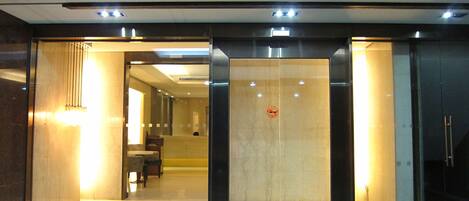 A modern hotel entrance with a glass door, a reception desk inside, and a staircase leading to the upper floor.