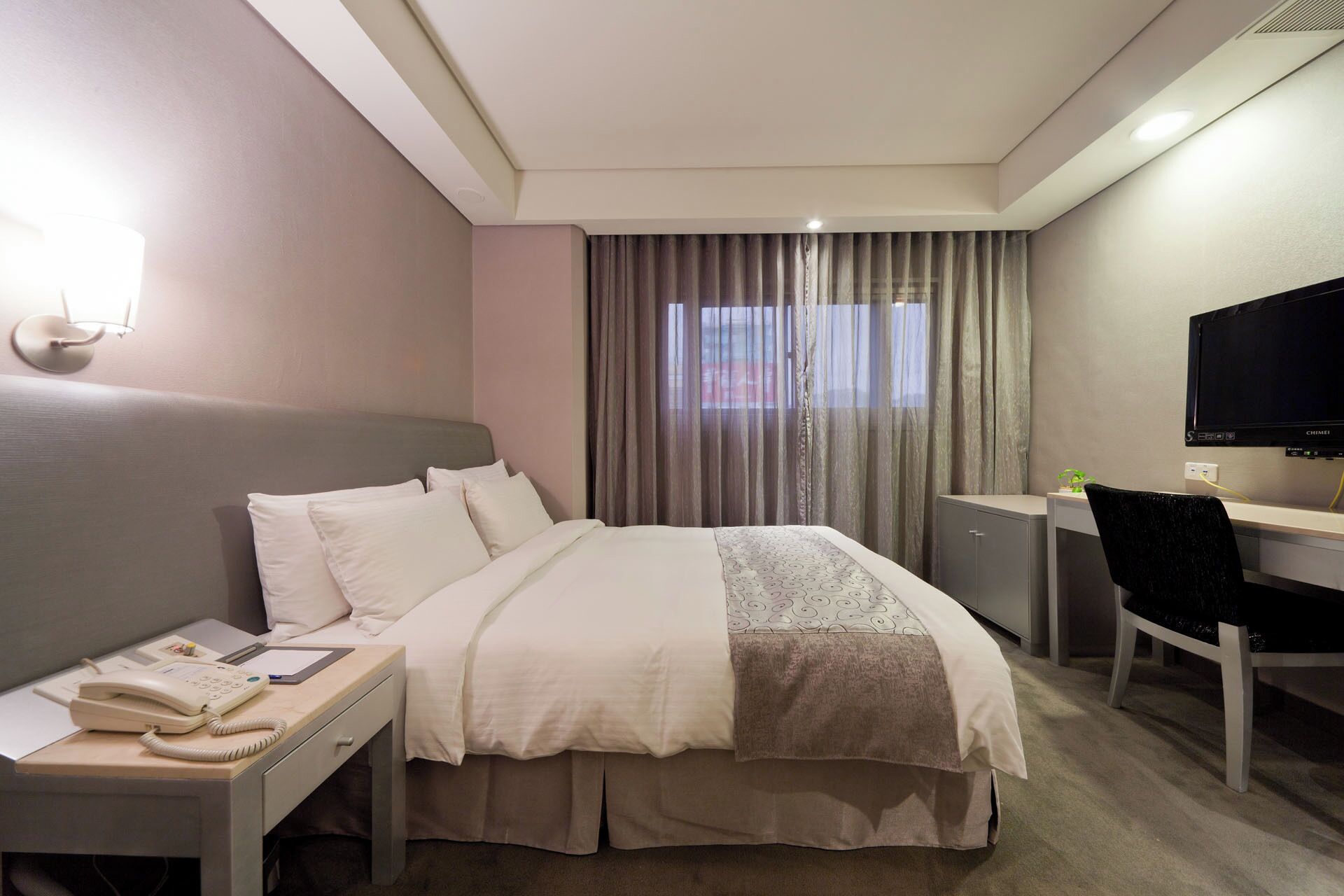 A hotel room with a bed, a desk with a chair, a telephone, and a television.