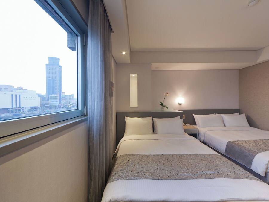 A hotel room with two beds, a window with a view of a cityscape, a bedside table with a lamp, and a wall-mounted light.