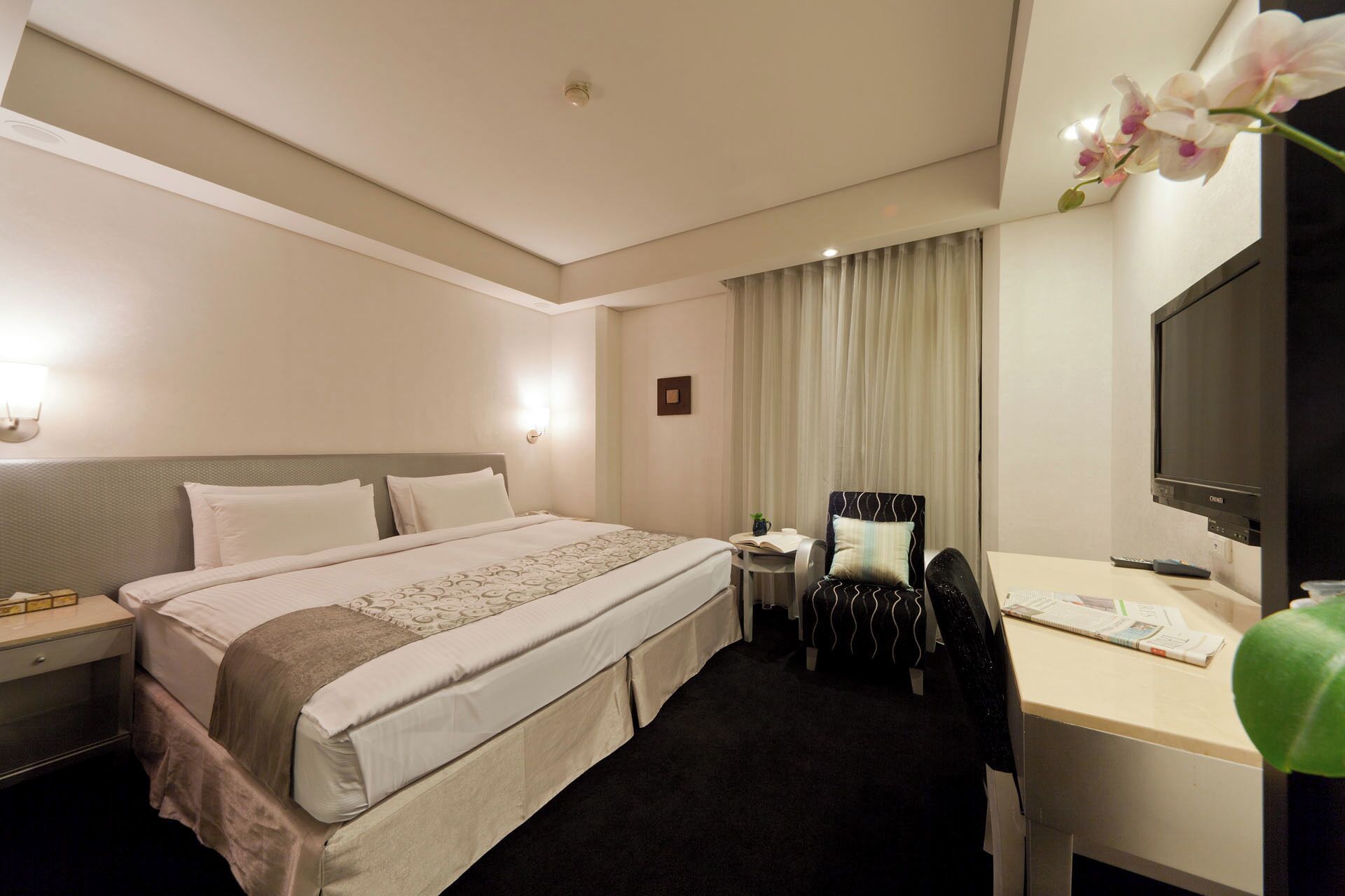 A hotel room with a large bed, a desk with a chair, a television, and a window with curtains.