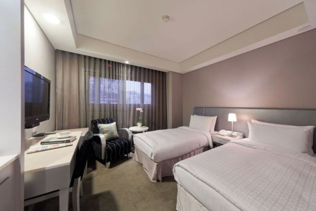 A hotel room with two beds, a desk, a TV, and a window with curtains.