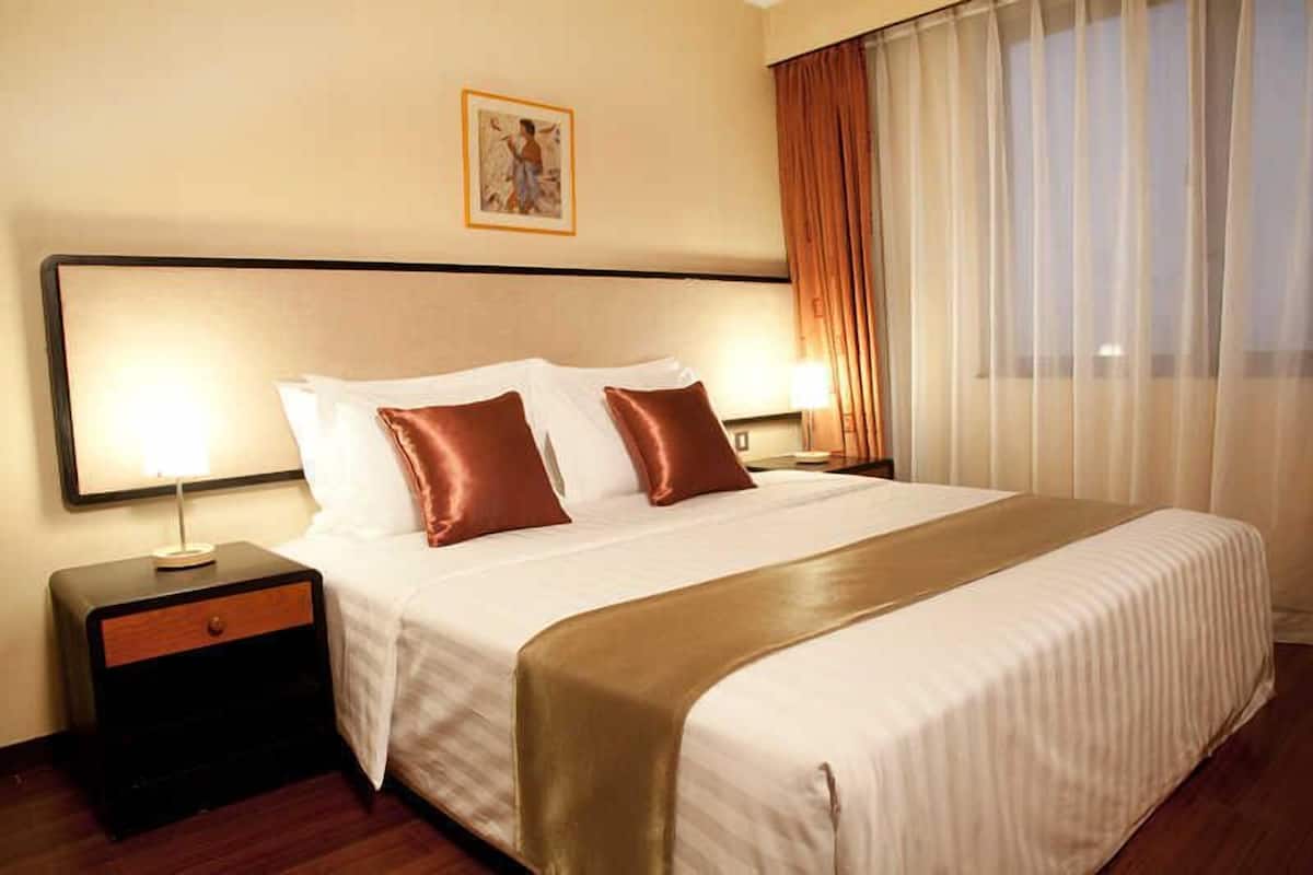 Une chambre d’hôtel avec un grand lit, deux tables de chevet, une commode, un tableau encadré au mur et une fenêtre avec des rideaux.