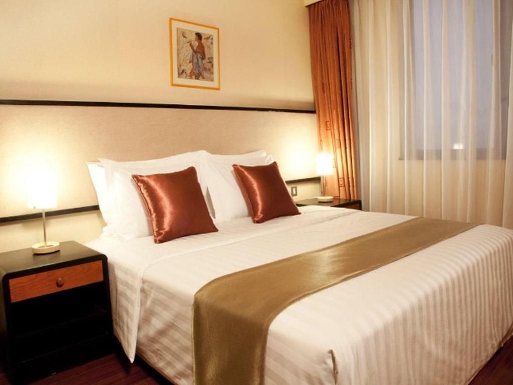 A hotel room with a large bed, two bedside tables, a nightstand, a framed picture on the wall, and a window with curtains.