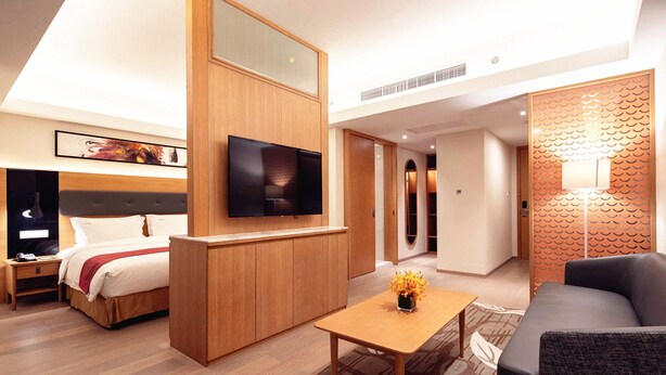 Quarto de hotel moderno com cama, televisão, secretária de madeira, sofá e um painel decorativo.