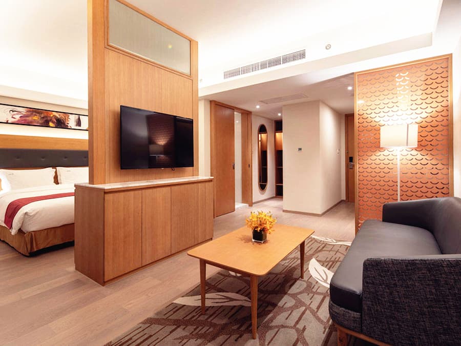 A modern hotel room with a bed, a TV, a wooden desk, a sofa, and a decorative wall.