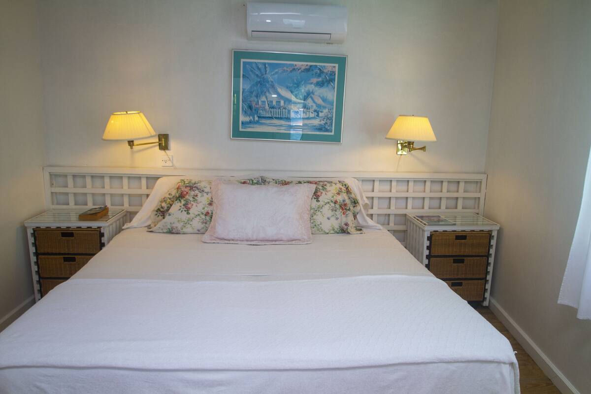 Een keurig opgemaakt bed met bloemenbedrukking, twee nachtkastjes met lampen, een wandairconditioner en een ingelijst beeld boven het bed.
