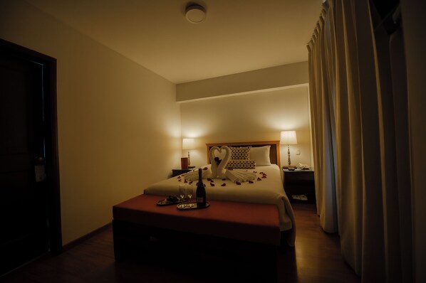 Quarto de hotel com cama, abajures, mesa de cabeceira com uma garrafa e copos, e uma porta.