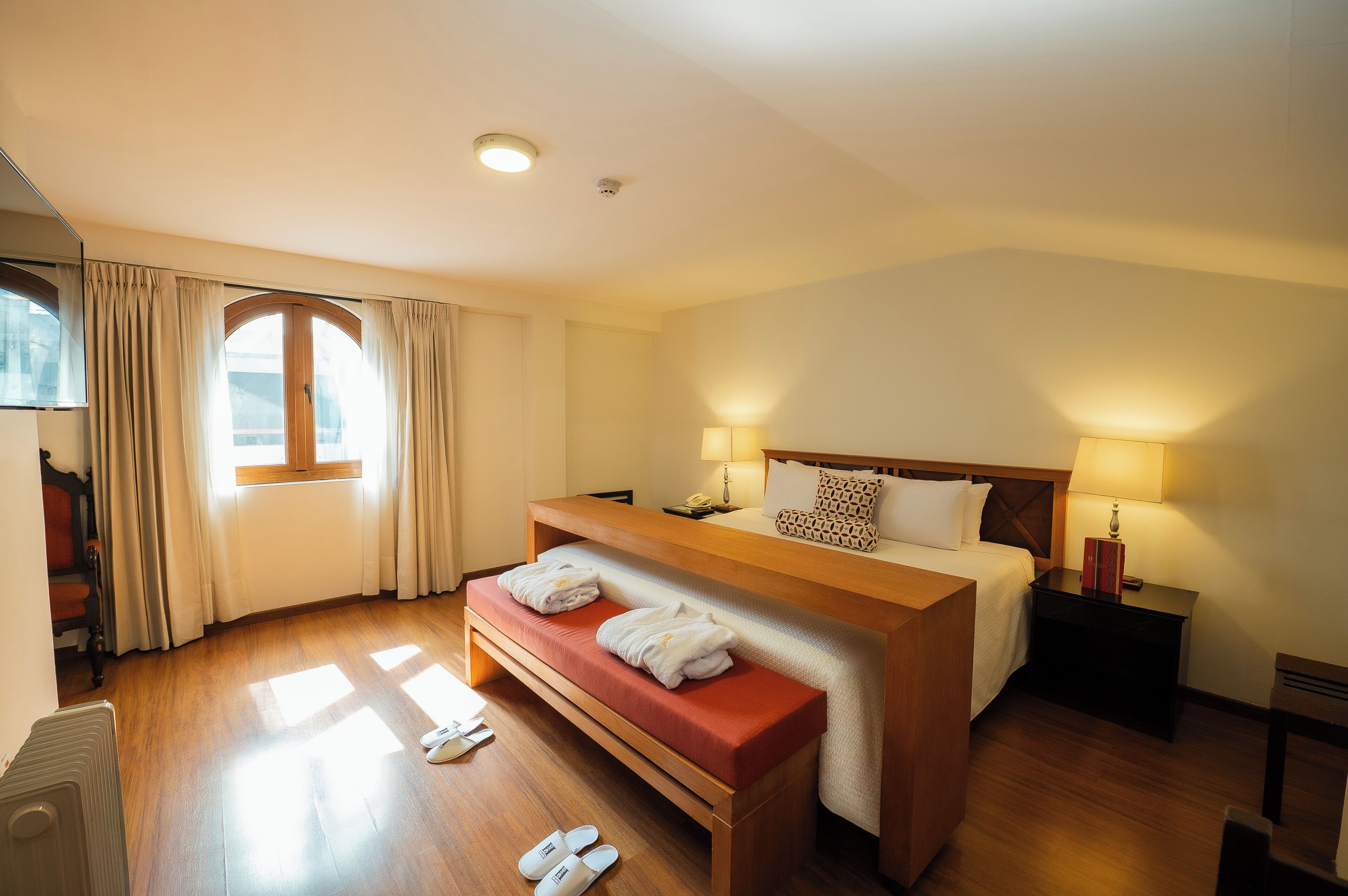 A hotel room with a wooden bed, a bench, a desk, and a window with curtains.